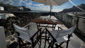 Una mesa con tapa de madera y un paraguas beige, ubicada en un patio de piedra con vistas a la costa y el mar, con un edificio a lo lejos.