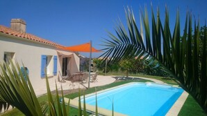 Una piscina con deck de baldosas, zona de estar techada al aire libre y follaje circundante.