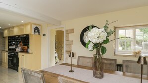 A dining area with a wooden table, chairs, and a vase with flowers. There is a kitchen in the background with a clock on the wall. - The Gate House (Cirencester)