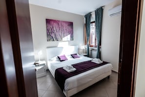 A hotel room with a bed, a nightstand, a lamp, a window with curtains, and a wall-mounted air conditioner. - DOMUS in PANTHEON (Rome)