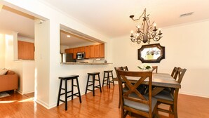 A dining area with a wooden table and chairs, a chandelier, and a kitchen in the background.