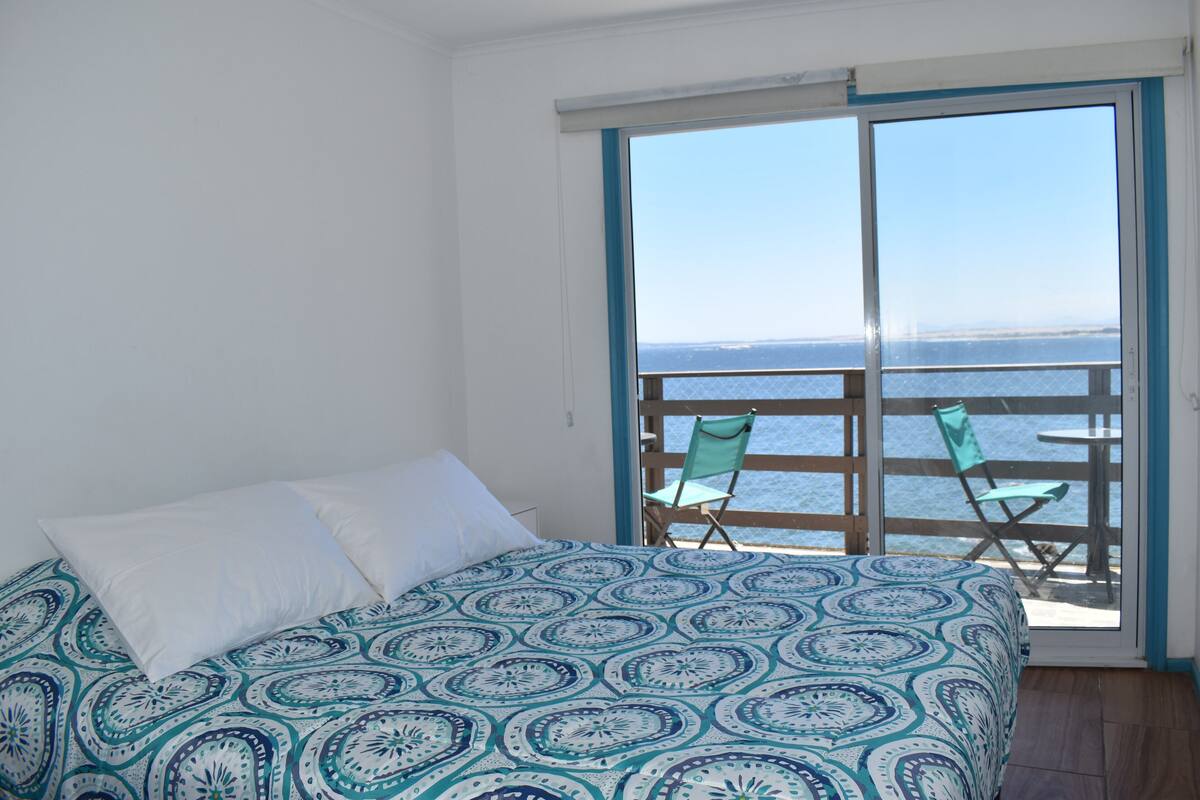 A bedroom with a bed, a window, and a view of the sea.