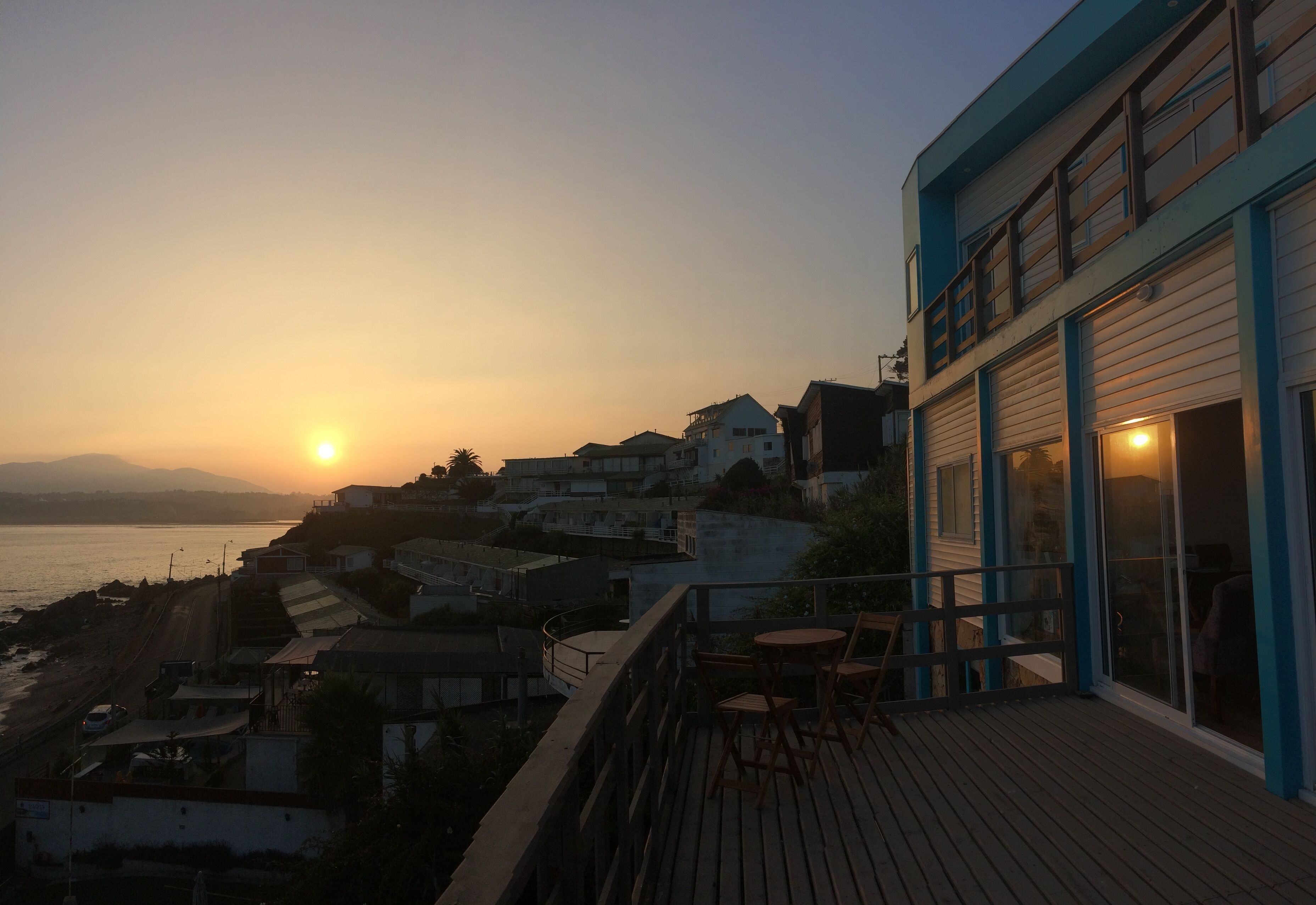 A coastal building with a deck overlooking the sea, featuring a view of the sunset and distant mountains.