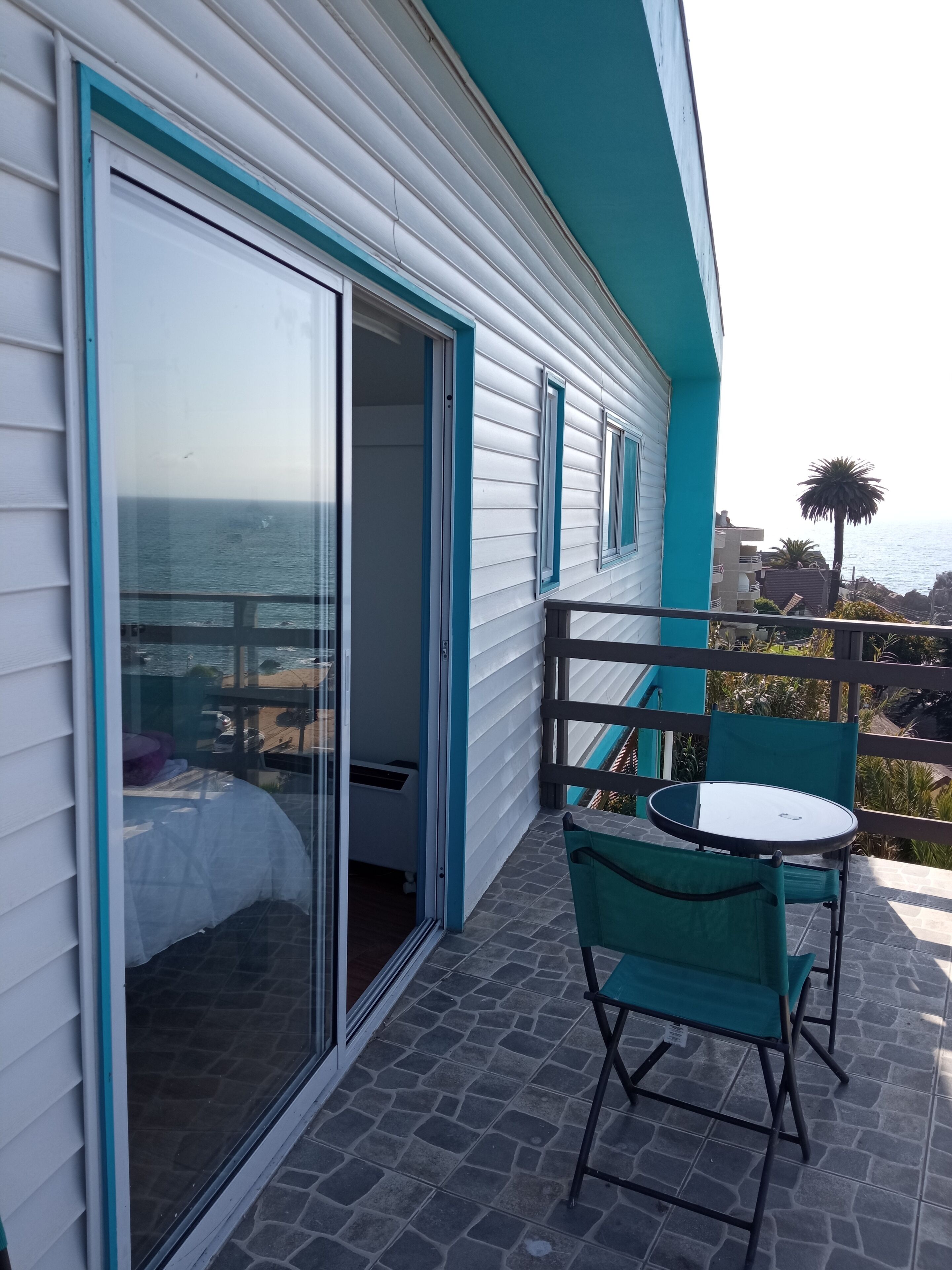 A balcony with a table and chairs, a sliding glass door leading to a bedroom, and a view of the ocean.