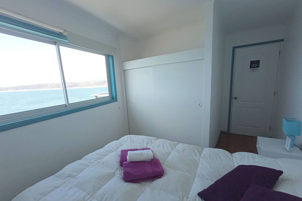 A bedroom with a bed, a window with a view of the sea, and a door.