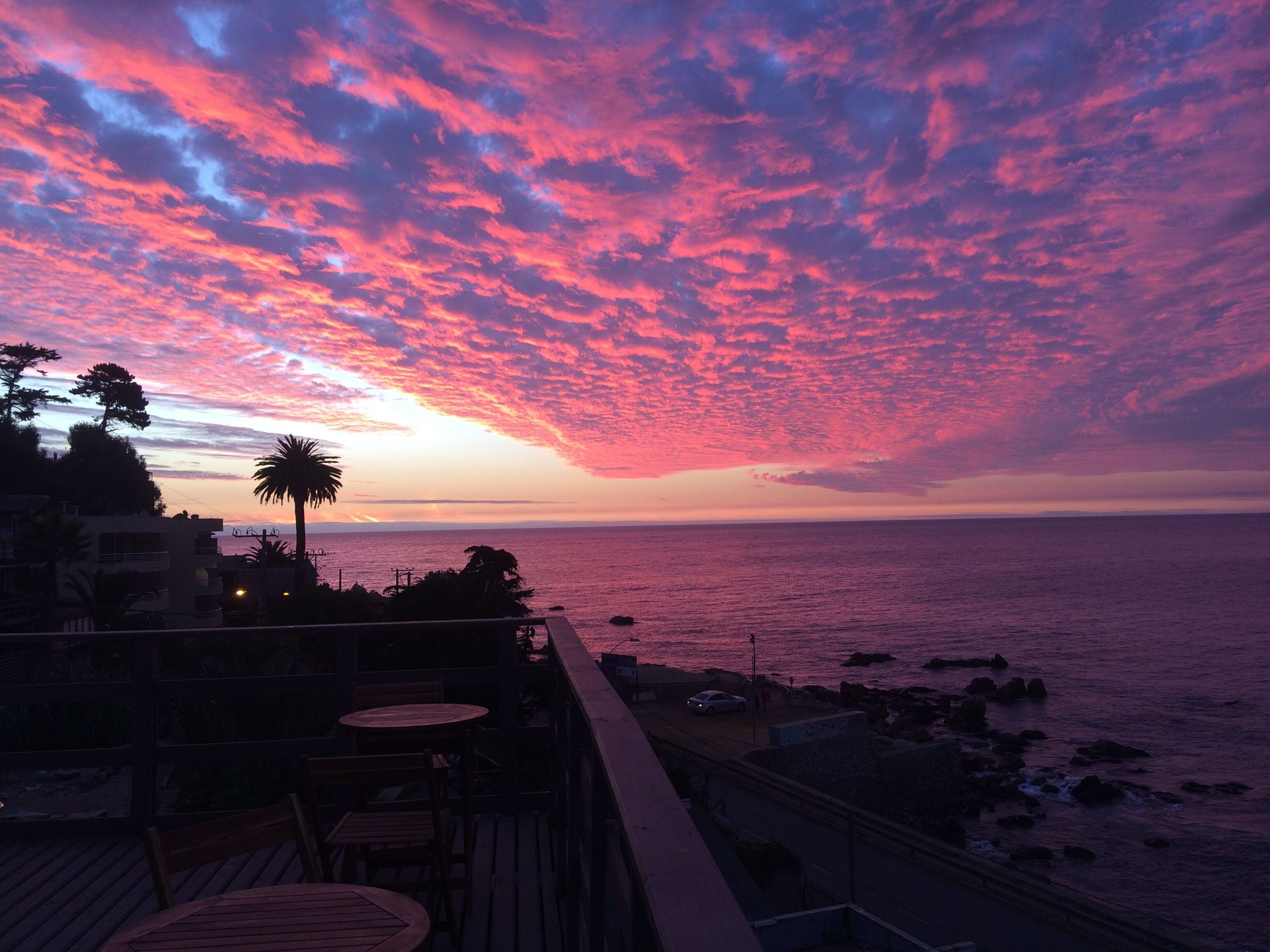 A sunset view from a balcony overlooking the sea, with a wooden deck and chairs.