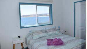 Un dormitorio con cama, mesita de noche, ventana y vista al mar, además de una puerta.