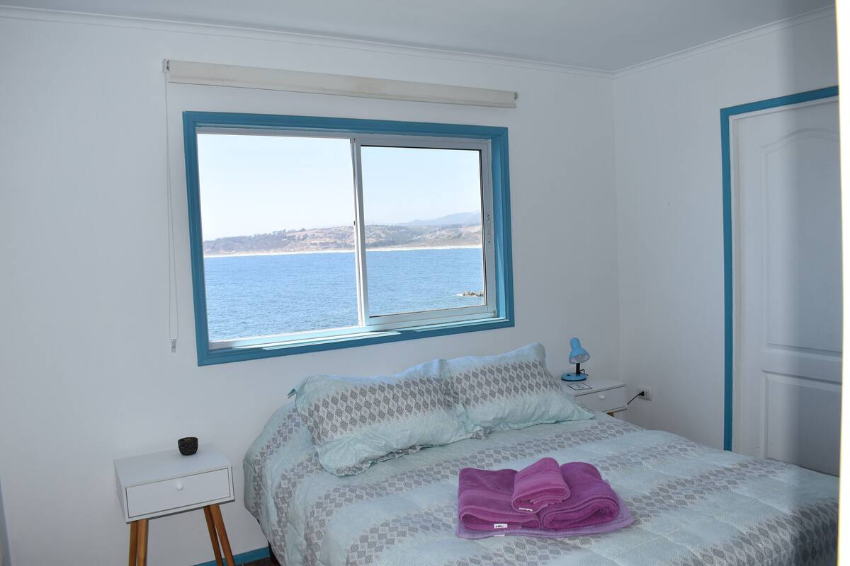 A bedroom with a bed, a nightstand, a window with a view of the sea, and a door.