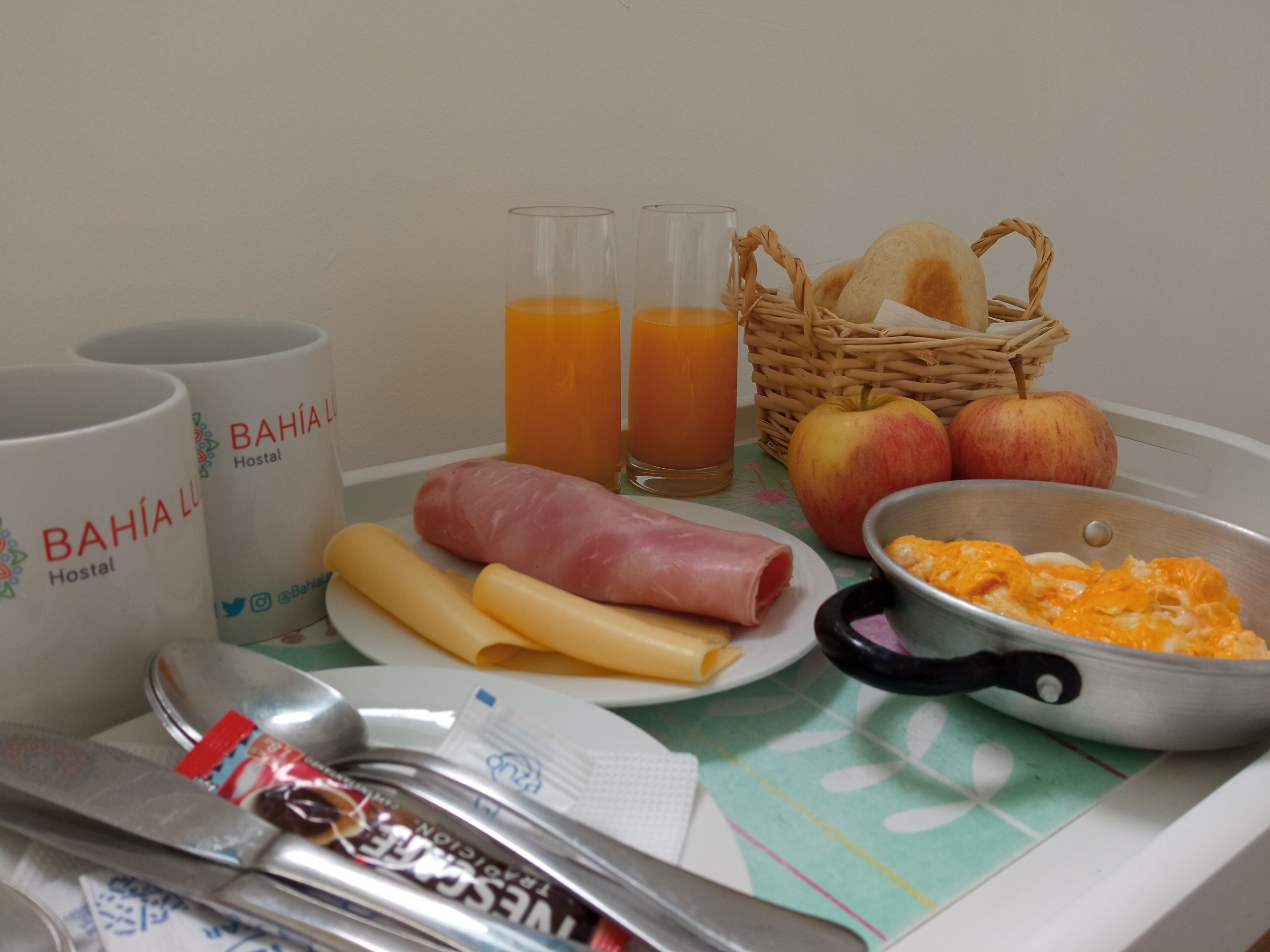A breakfast setup with two cups labeled "BAHIA LUZ Hostel", a plate with ham and cheese, a pan with scrambled eggs, a basket with bread, and two glasses of orange juice.