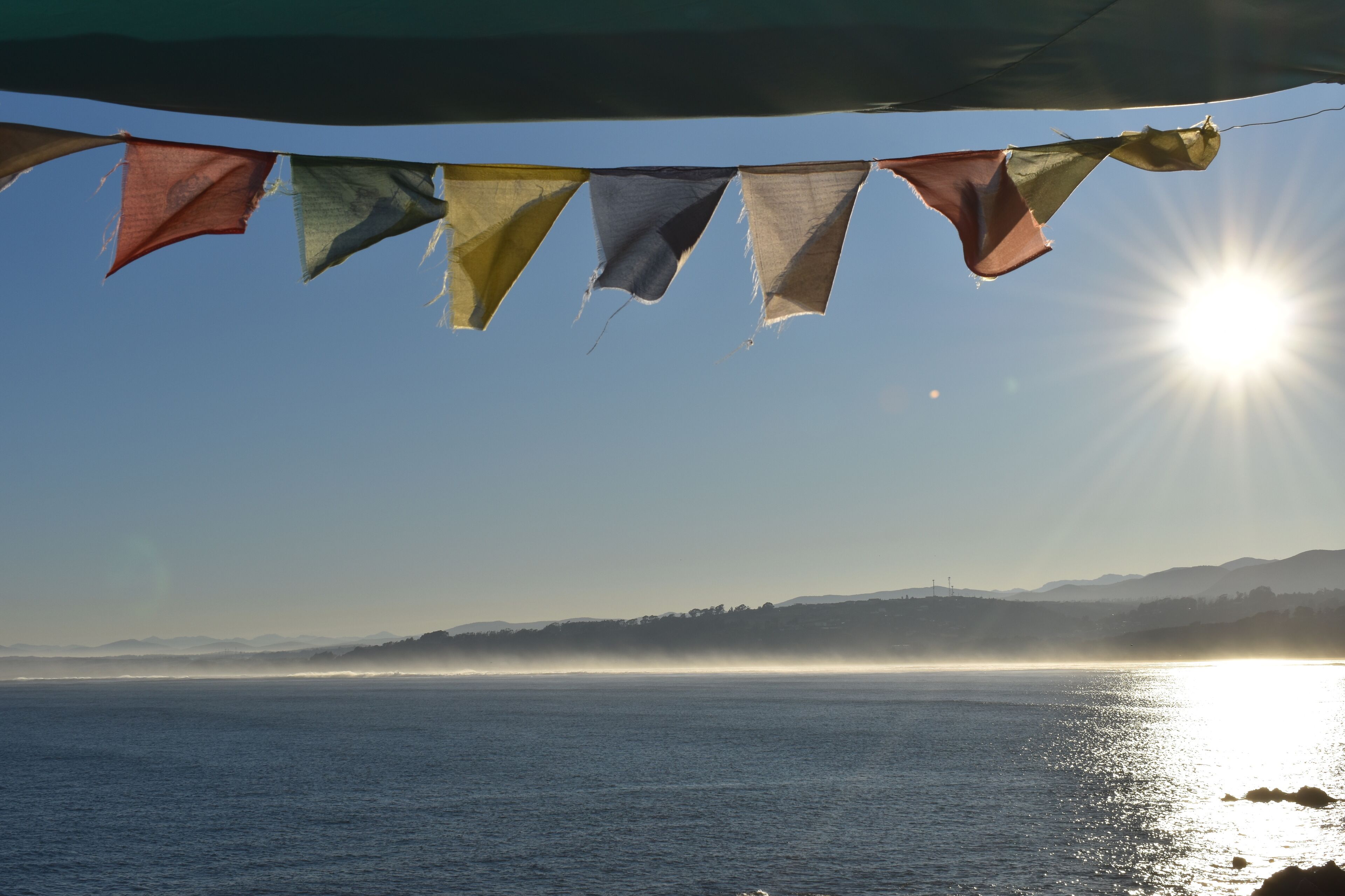 Uitzicht op de kust onder een heldere hemel, met de zon die in het water weerkaatst, en een rij vlaggen die wapperen.
