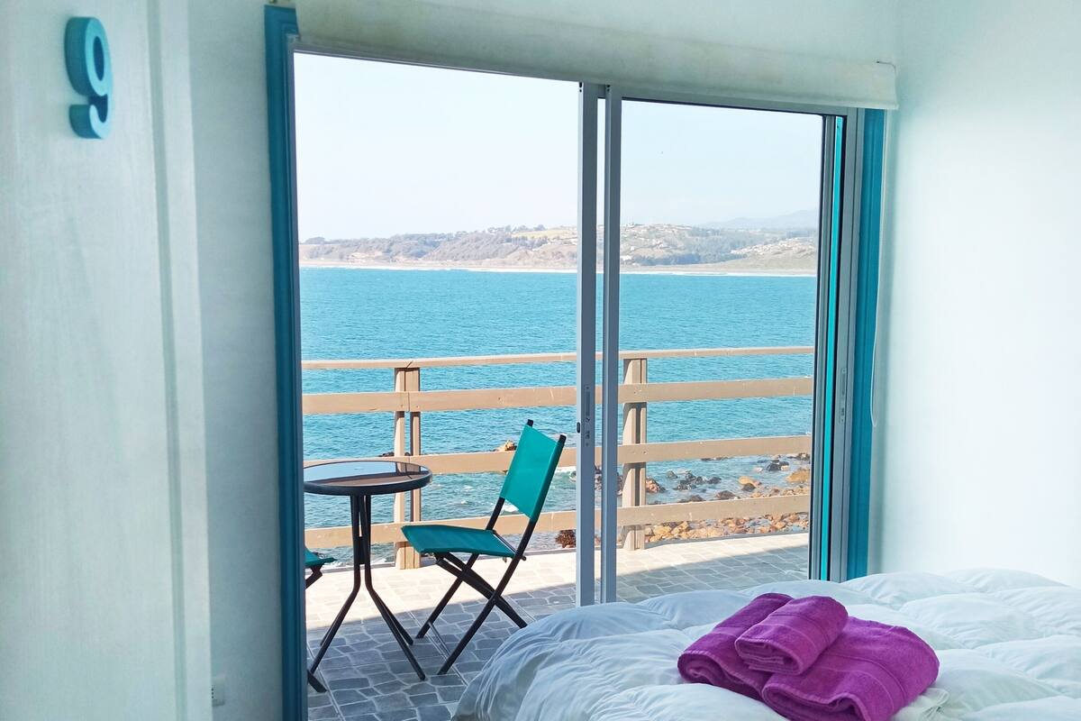 A bedroom with a bed, a chair, and a table by a large window overlooking a beach.
