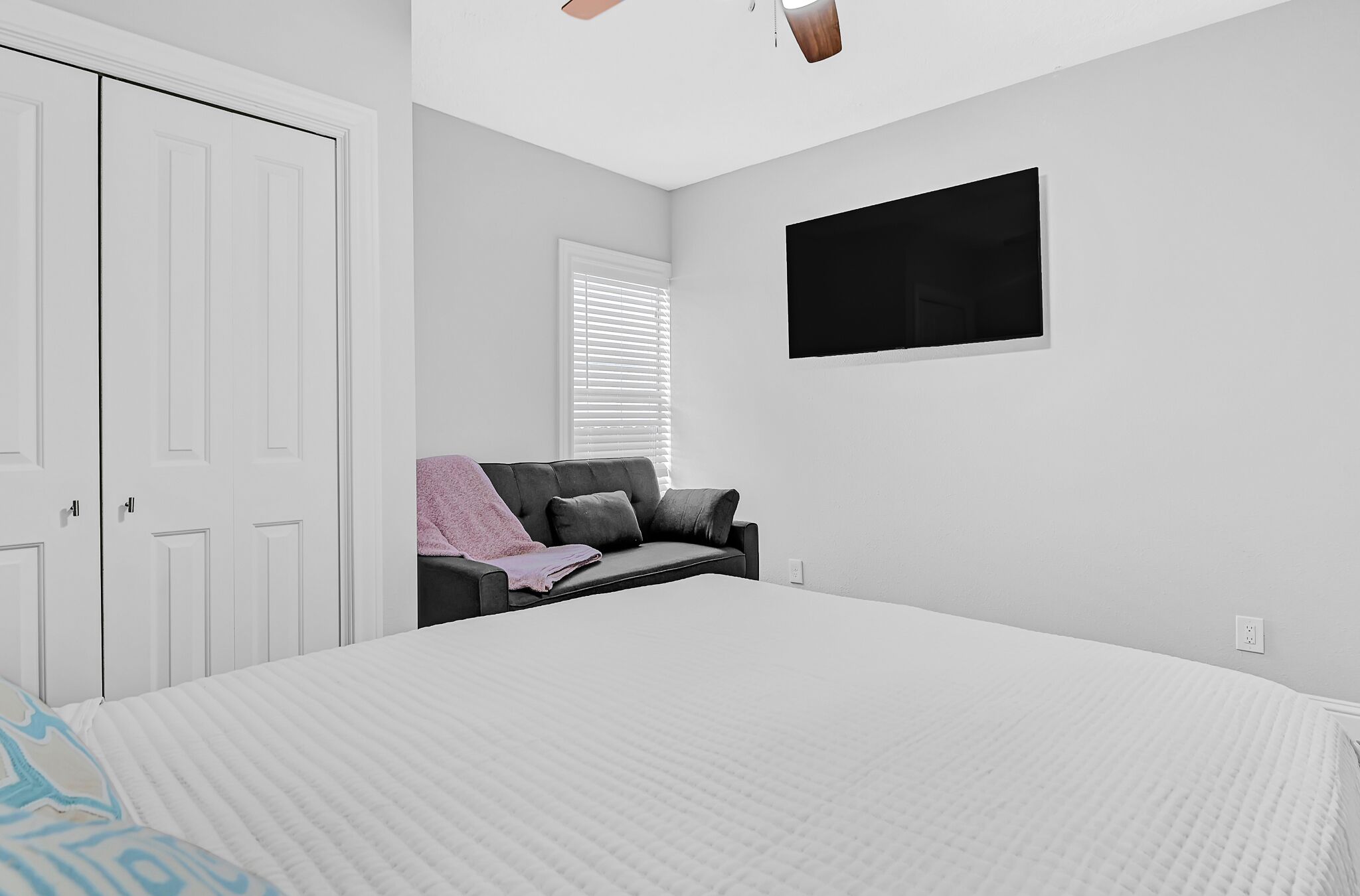 A bedroom with a large bed, a grey sofa, a TV mounted on the wall, and white wardrobes.