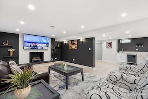 Ein modernes Wohnzimmer mit Flachbildfernseher, Kamin, einer Couch und einer Küche mit weißen Schränken.