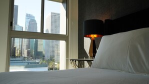 Une chambre d’hôtel dotée d’une grande fenêtre donnant sur la ville, d’un lit avec du linge de maison blanc et d’une lampe de table avec un abat-jour noir.