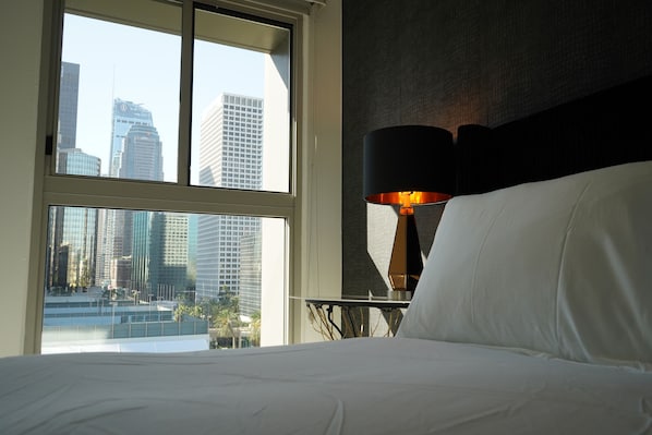 A hotel room with a large window offering a city view, a bed with white linens, and a table lamp with a black shade.