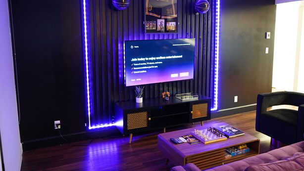 Un salon moderne avec un téléviseur à écran plat fixé à un mur foncé, une table basse en bois sur laquelle est posé un jeu d’échecs, et un canapé violet.