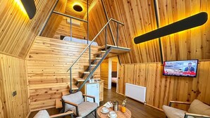Ein gemütliches Blockhaus-Interieur mit Holzvertäfelung, einer Treppe, einer Essecke mit Tisch und Stühlen sowie einem an der Wand befestigten Fernseher.