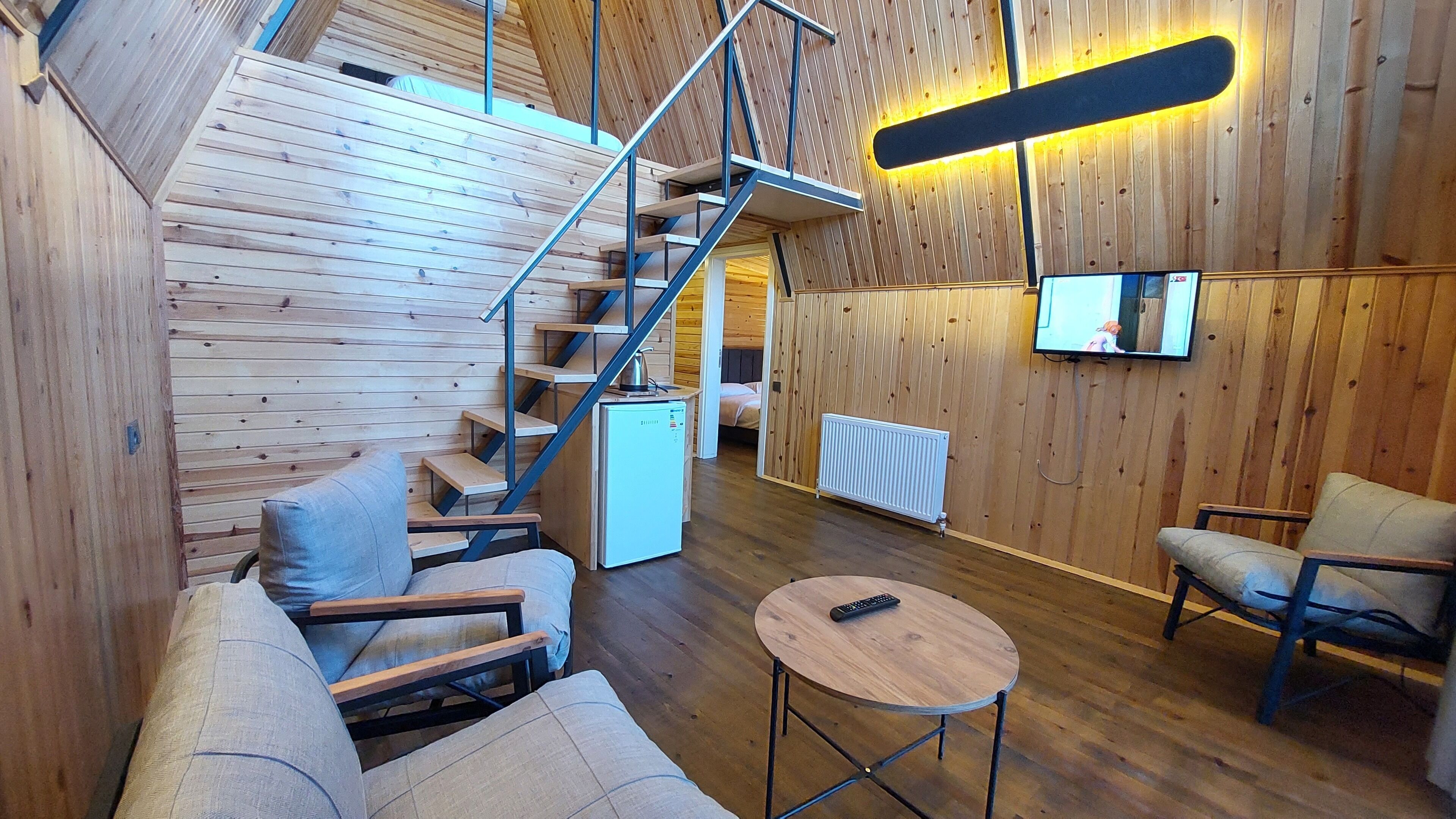 A cozy cabin interior with a wooden staircase, a small kitchenette, a sofa, a round table, and a TV mounted on the wall.
