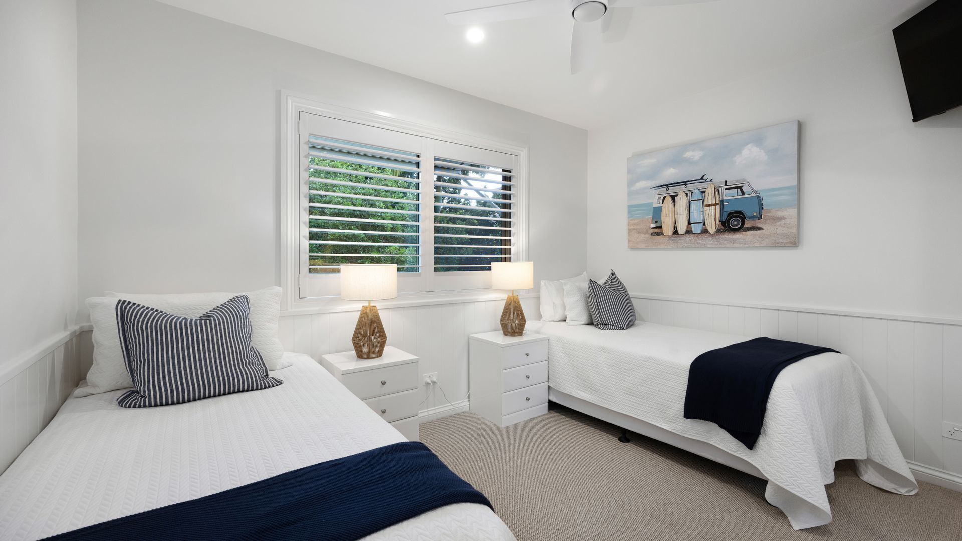 A bedroom with two beds, a large window with blinds, bedside tables with lamps, a wall-mounted TV, and a beach-themed painting.