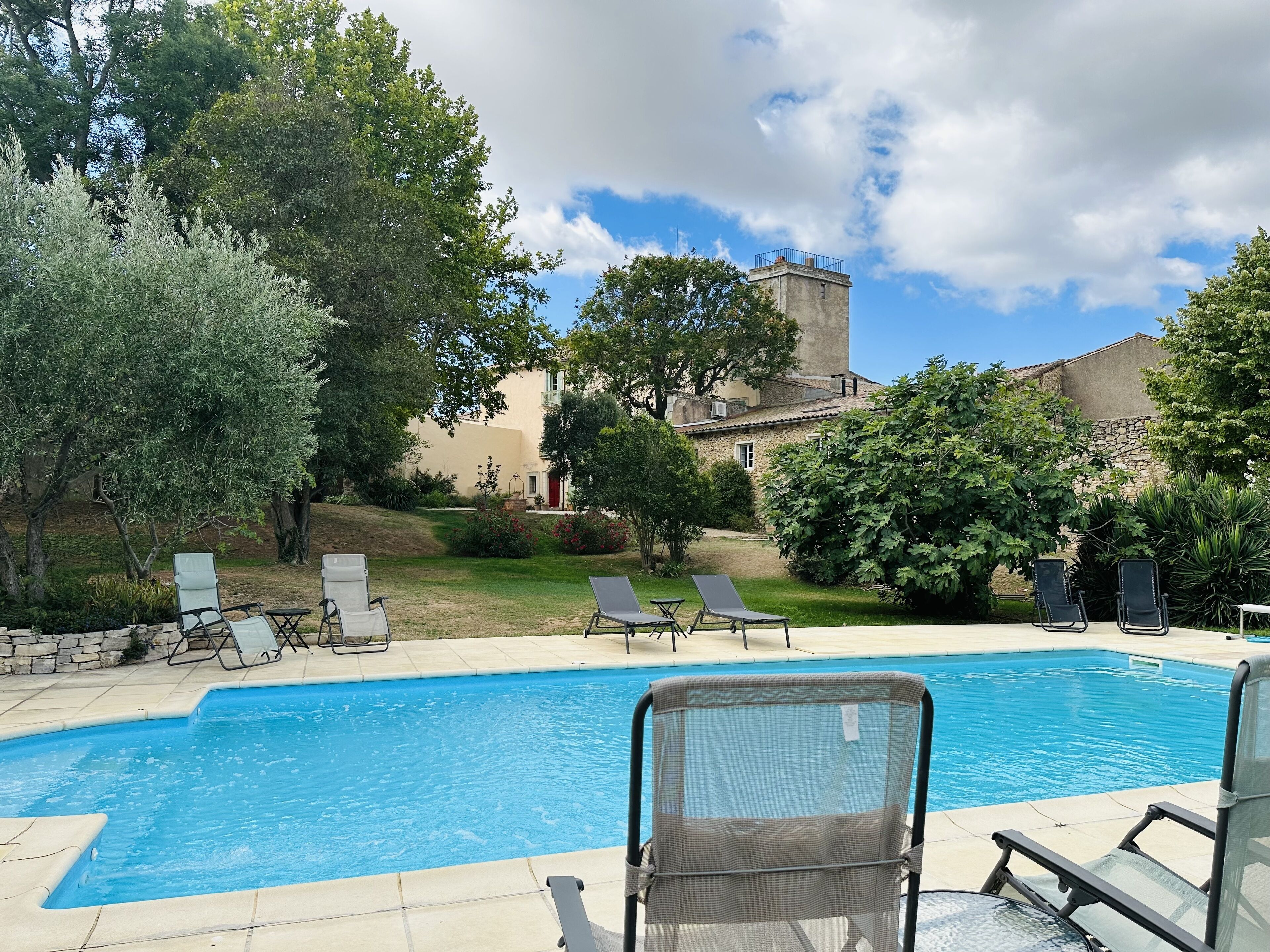 A swimming pool with lounge chairs, surrounded by greenery and a stone building in the background.