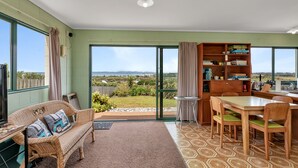 Una habitación espaciosa con un gran ventanal corredera que da a un balcón, un sofá de mimbre con cojines, una mesa de comedor de madera con sillas y una estantería llena de libros y objetos decorativos.