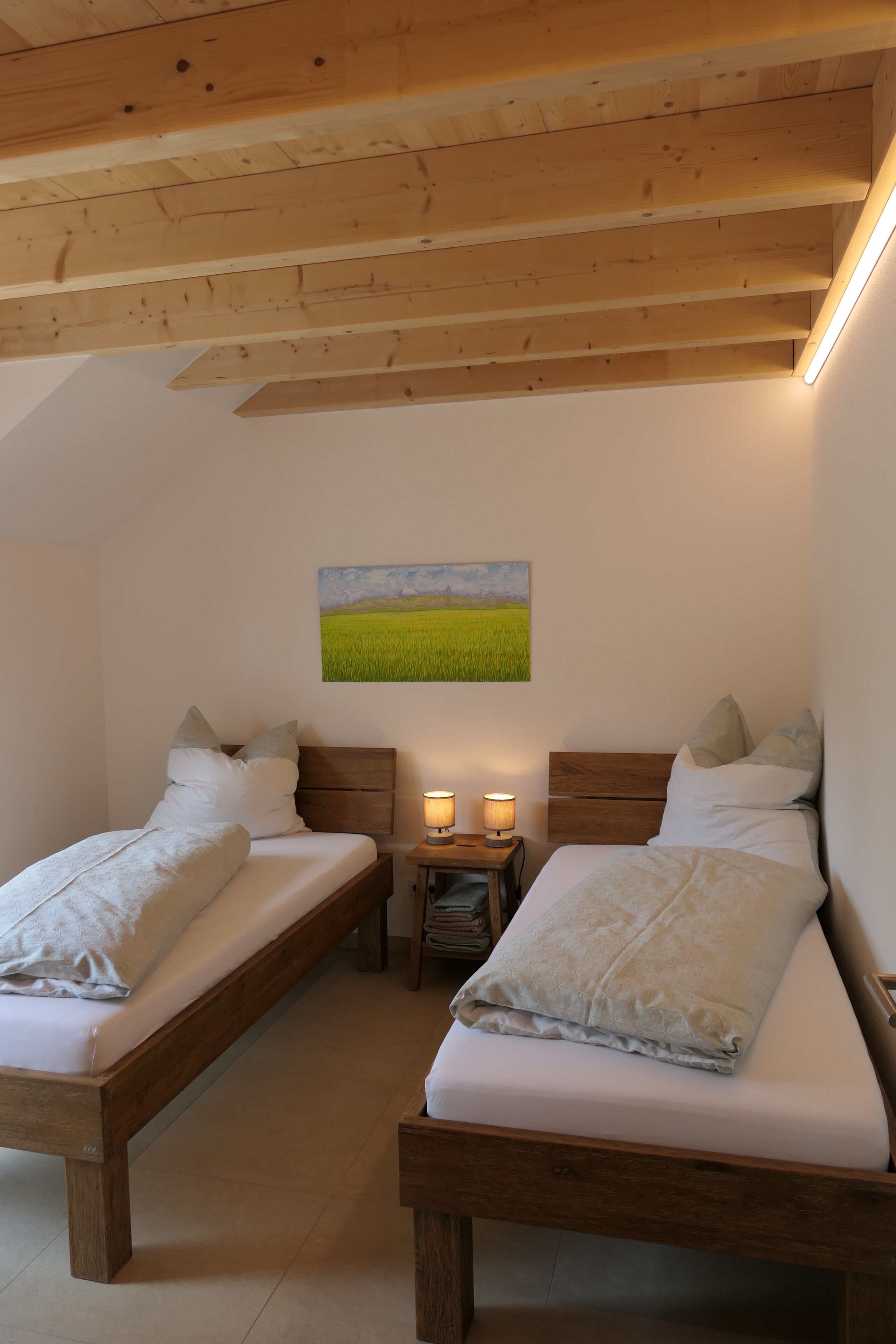 Une chambre avec deux lits en bois, une table de chevet avec deux lampes et un tableau représentant un champ accroché au mur.