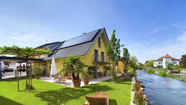 Una casa amarilla con techo de paneles solares, un río y un césped con muebles de exterior.