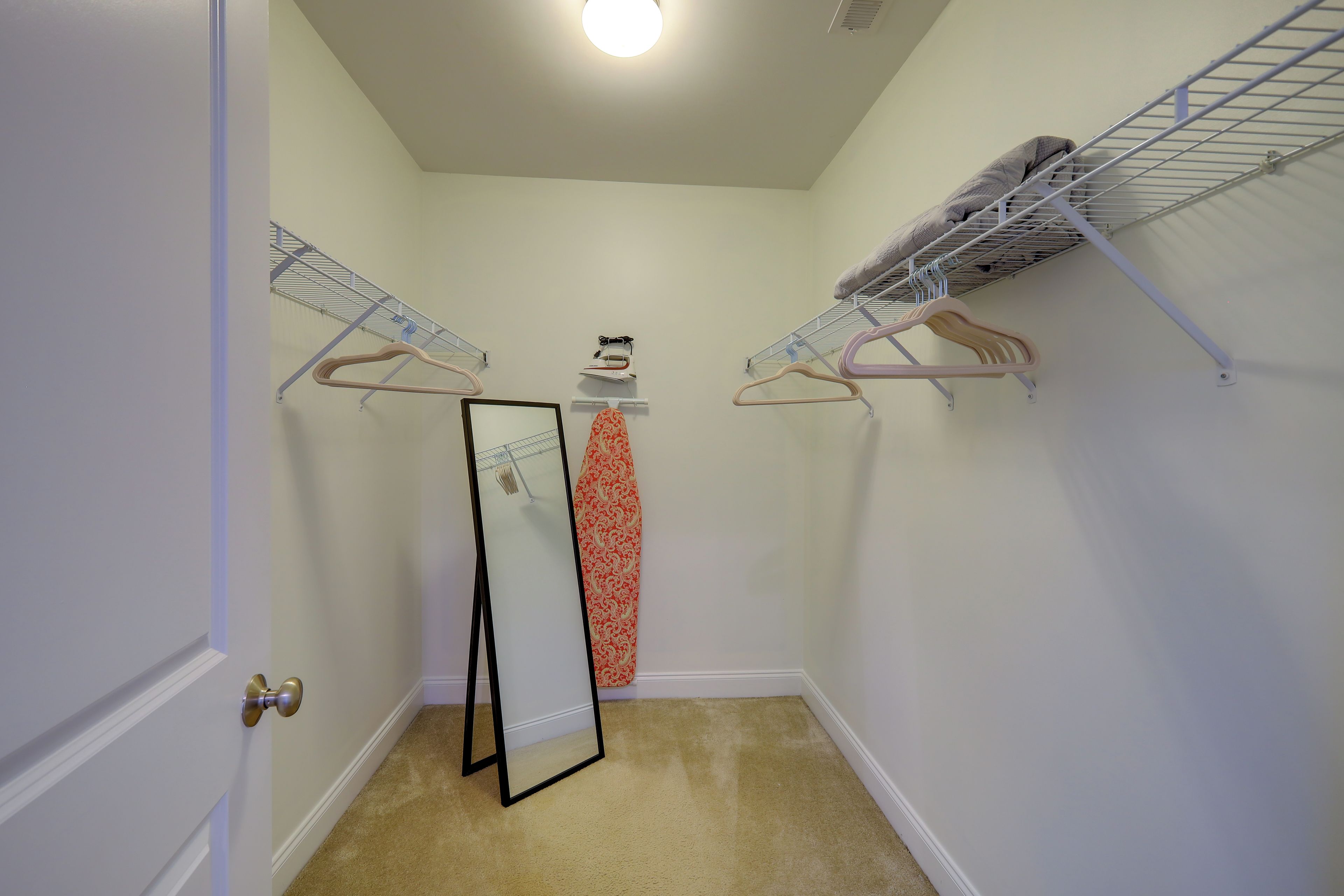 A small, carpeted closet with two metal drying racks, a mirror, and a wall-mounted ironing board.