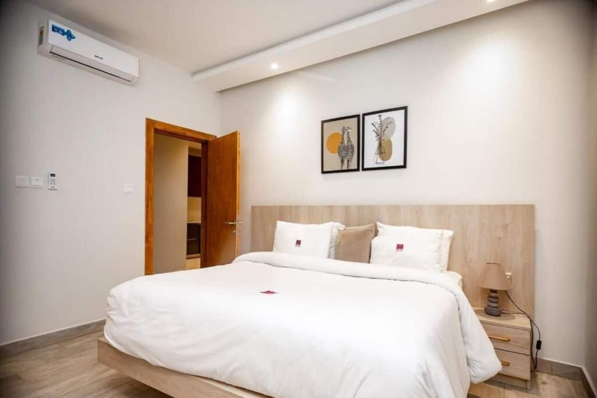 Un dormitorio moderno con estructura de cama de madera, ropa de cama blanca y una mesita de noche con lámpara.