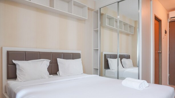 A neatly made bed with white linens and a brown headboard, a large mirror reflecting the room, and a wooden door to the right. - Good Deal And Cozy Stay Studio Room Tifolia Apartment (Jakarta)