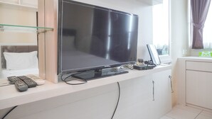 A modern living room with a large flat-screen TV mounted on a white wall, a glass shelf, and a white cabinet below. - Good Deal And Cozy Stay Studio Room Tifolia Apartment (Jakarta)