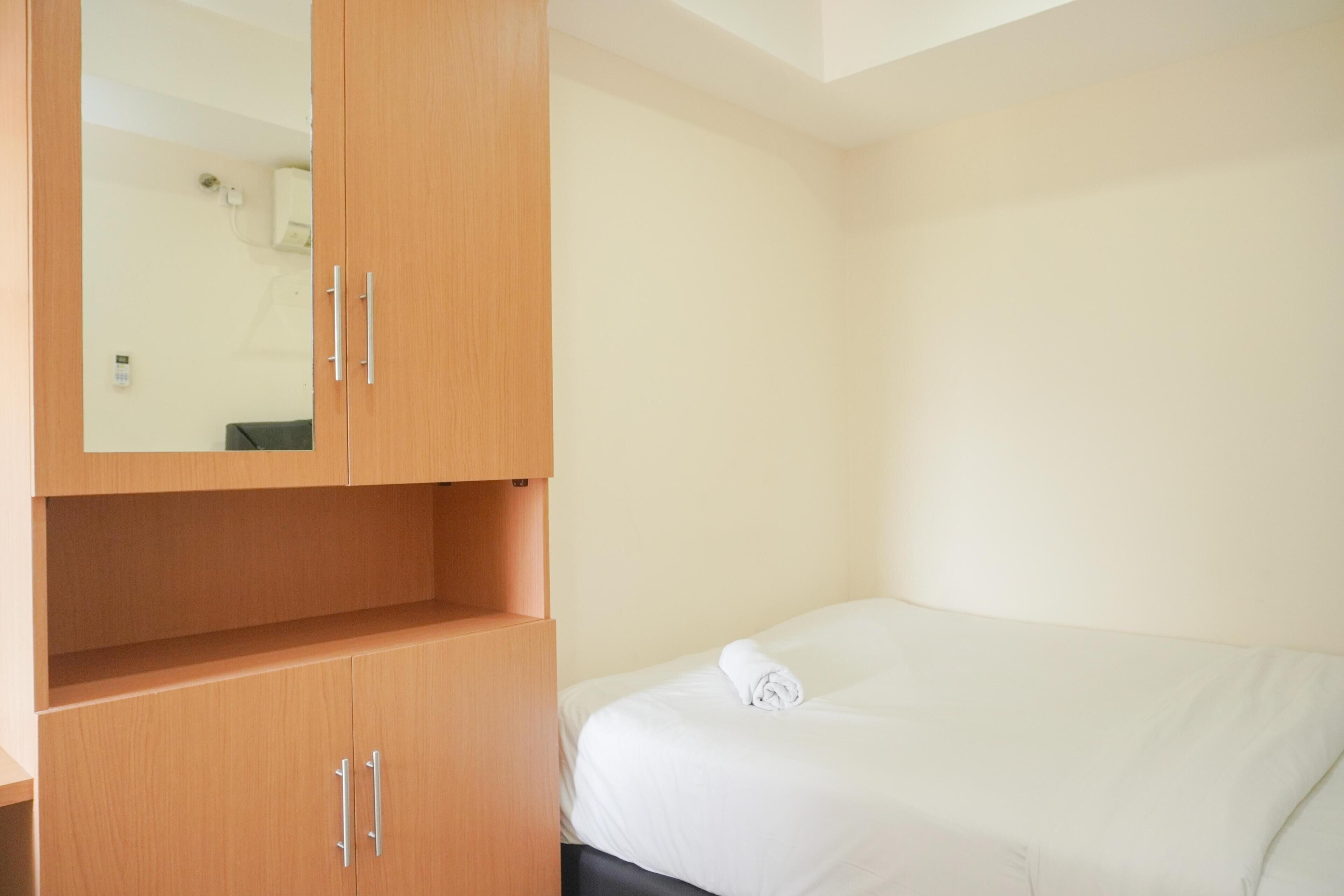 Una cama individual con colchón blanco y una toalla doblada cuidadosamente. Se observa un armario de madera con dos puertas y un espejo.