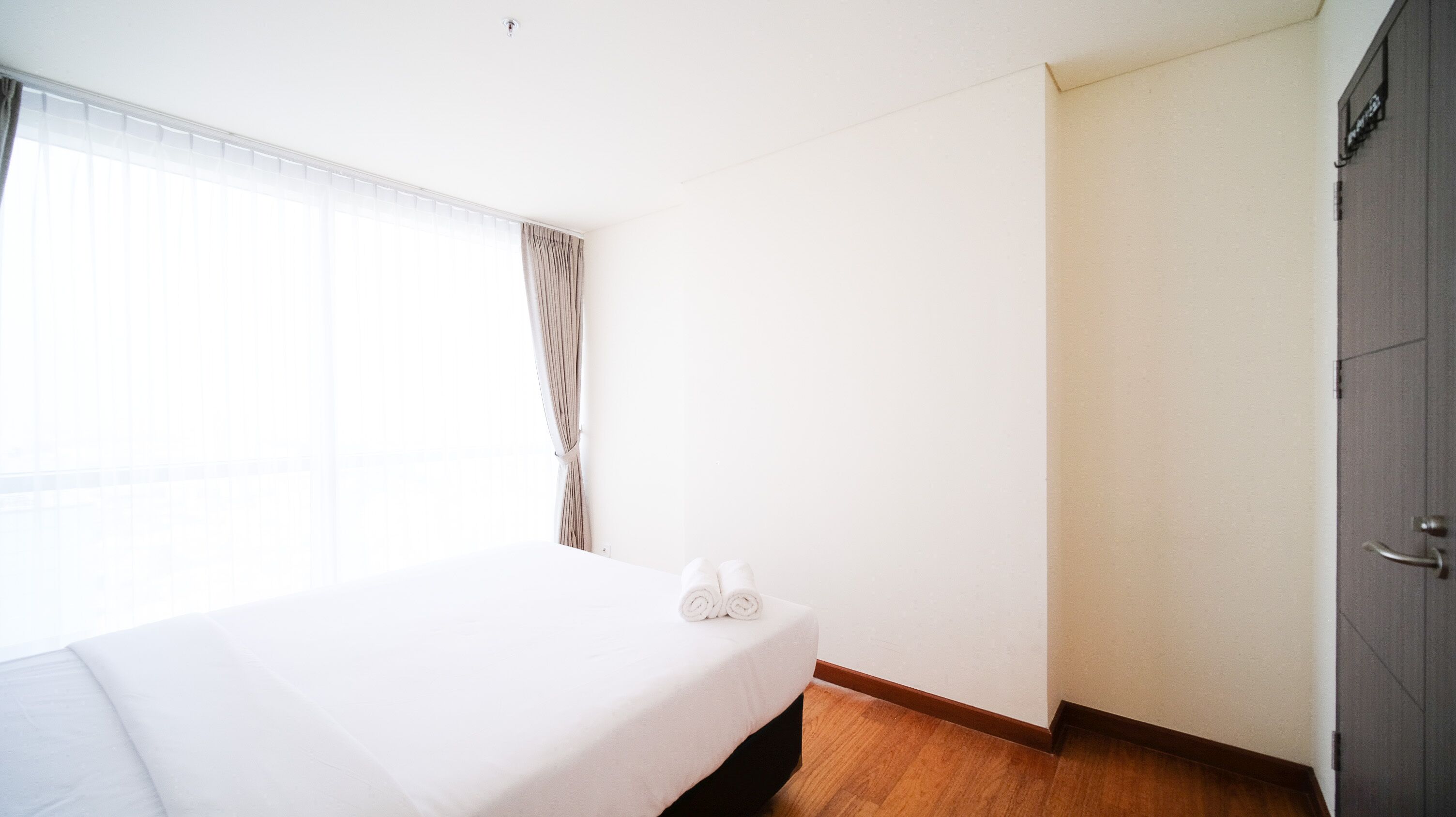 A hotel room with a bed, a window with curtains, a wooden floor, and a door.