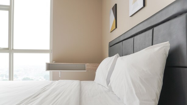 A neatly made bed with white linens in a room with a large window, a wall-mounted shelf, and a framed picture.