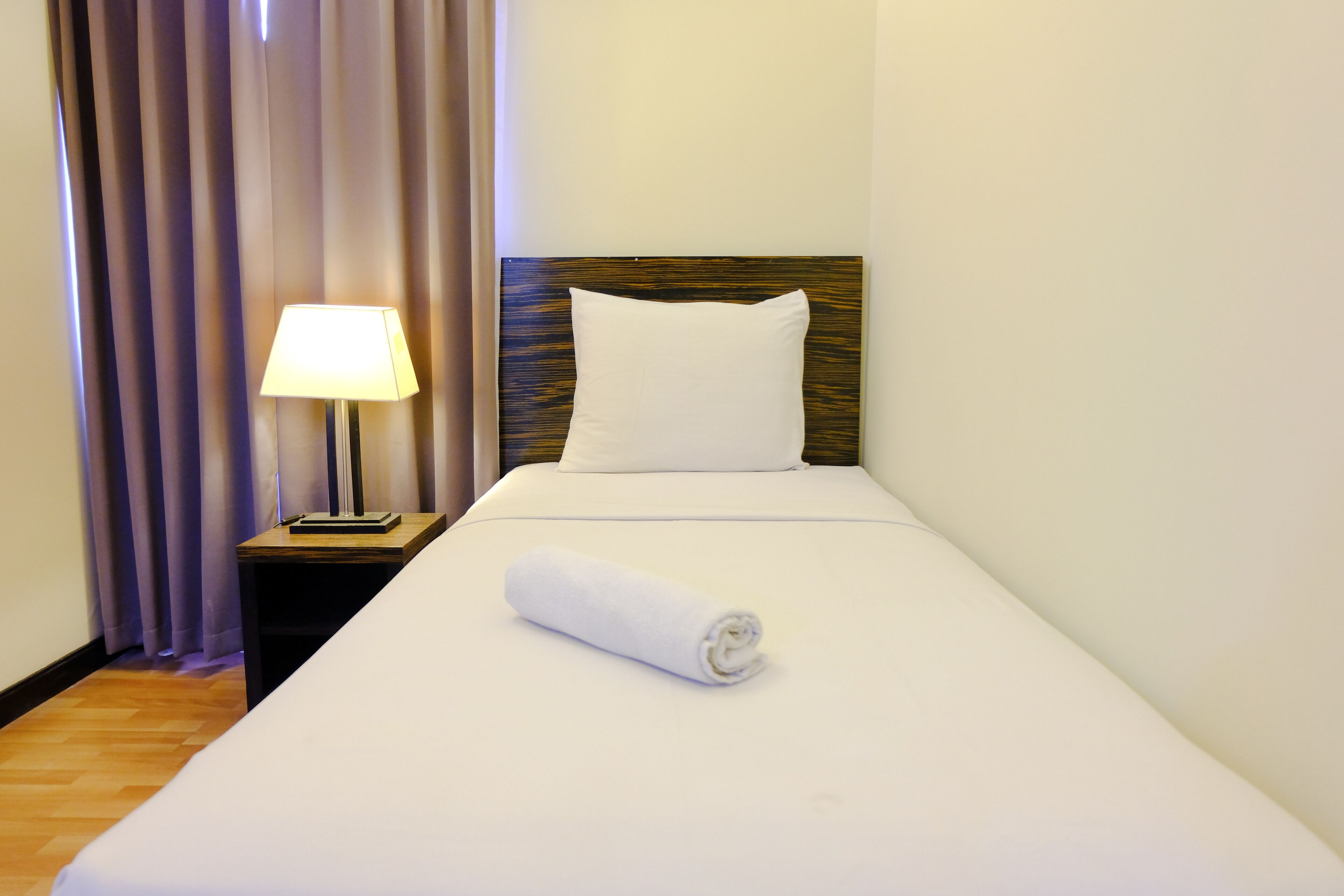 A neatly made bed with a rolled towel, a bedside lamp, and a curtain.
