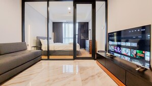 A modern hotel room with a large flat-screen TV displaying various streaming services, a grey sofa, and a bed with a white headboard. - Cozy And Elegant 1Br The Smith Alam Sutera Apartment (Tangerang)
