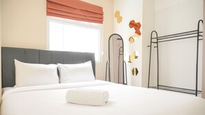A neatly made bed with white linens and a rolled white towel, a window with orange blinds, and a metal drying rack.