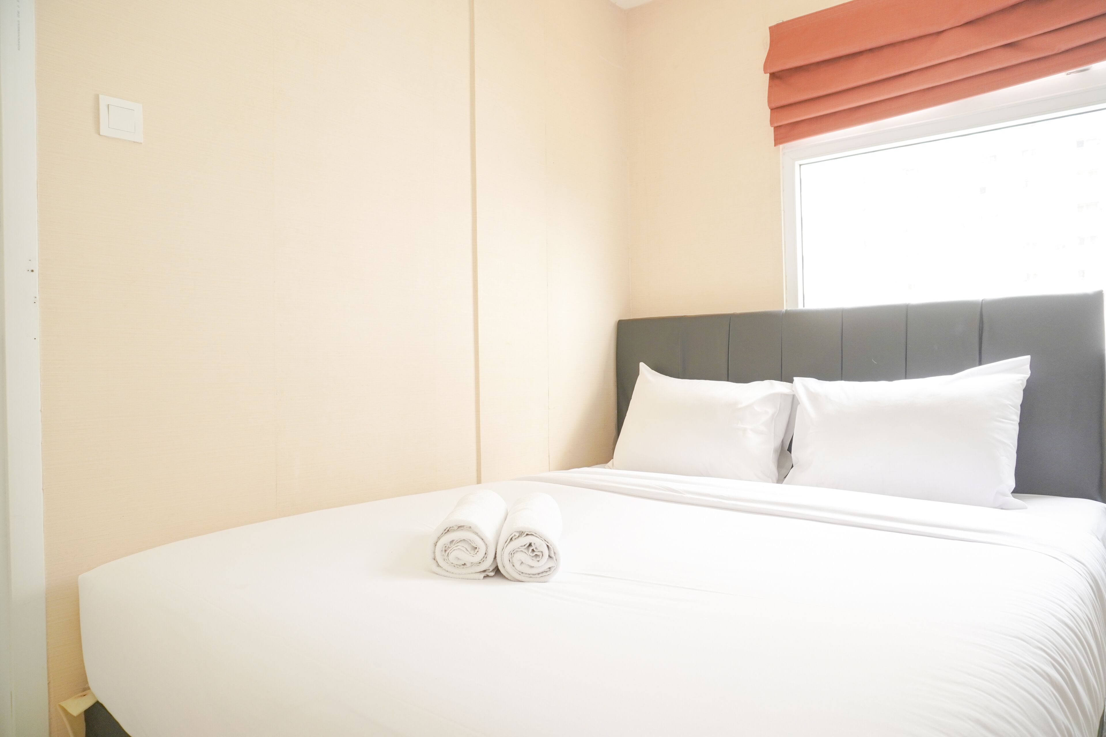 A neatly made bed with white linens and rolled towels, a grey headboard, and a window with a brown valance.