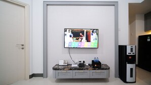 A hotel room with a flat-screen TV mounted on the wall, a marble countertop with a coffee machine, and a refrigerator. - Compact And Homey 2Br At Benson Supermall Mansion Apartment (Surabaya)