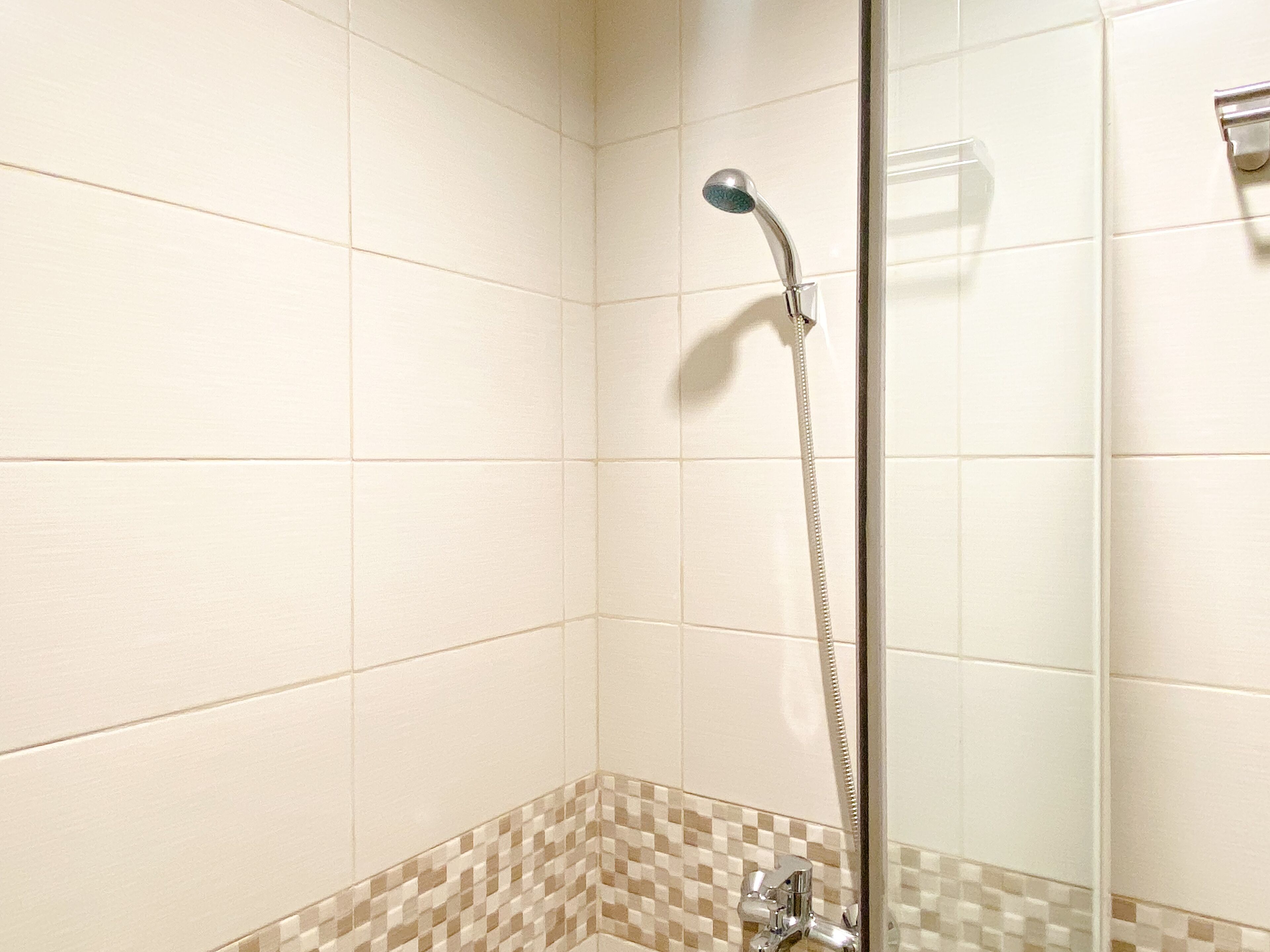 A shower with a glass door, white tiles, and a checkered tile border at the bottom.