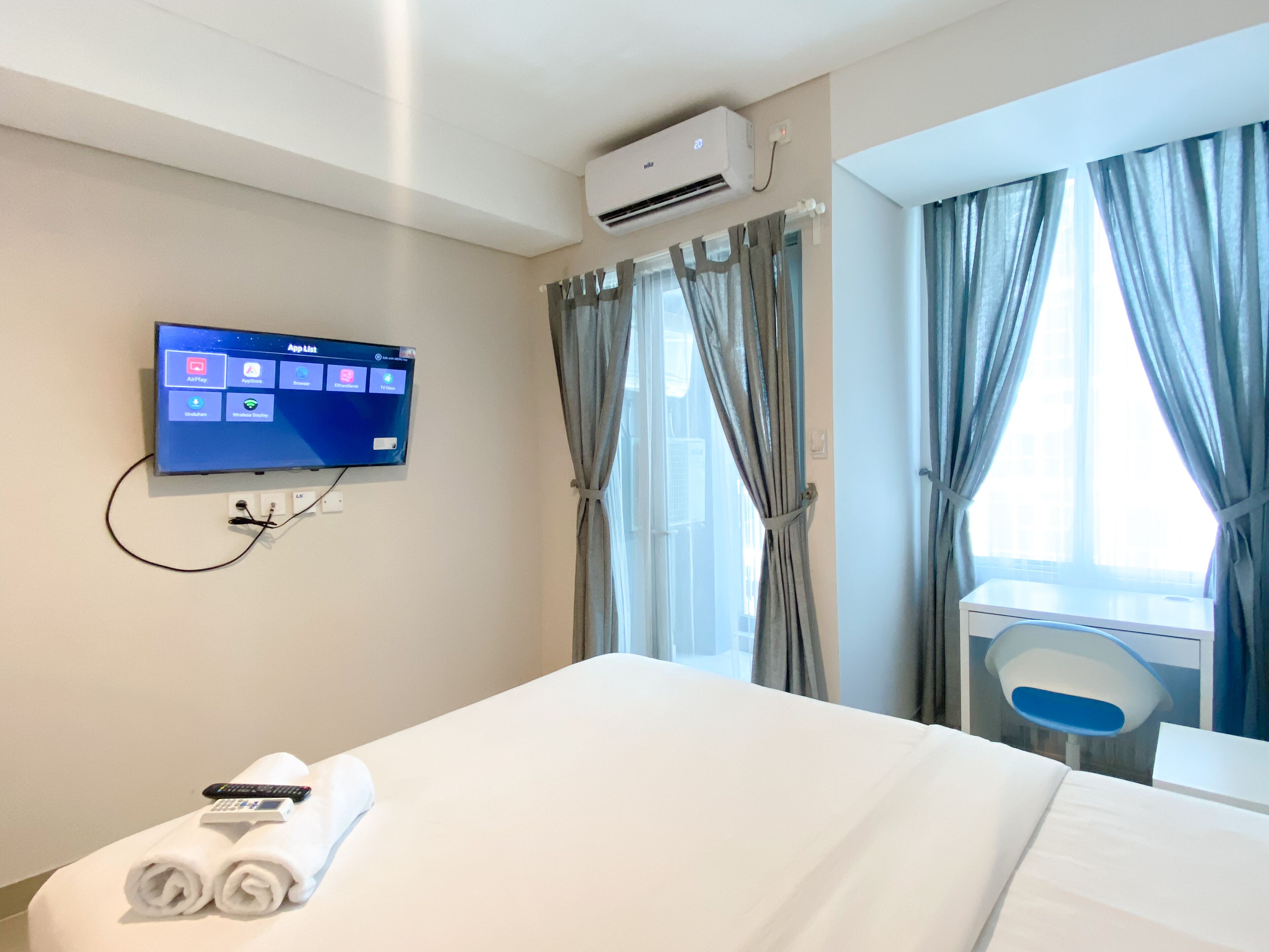 A hotel room with a bed, a TV mounted on the wall displaying an app list, a desk with a chair, and a window with curtains.