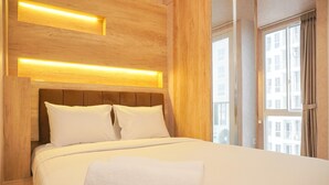 A neatly made bed with white linens and pillows, a wooden headboard, and a bedside table with a white towel. - Homey And Well Furnished Studio Tokyo Riverside Pik 2 Apartment (Teluknaga)