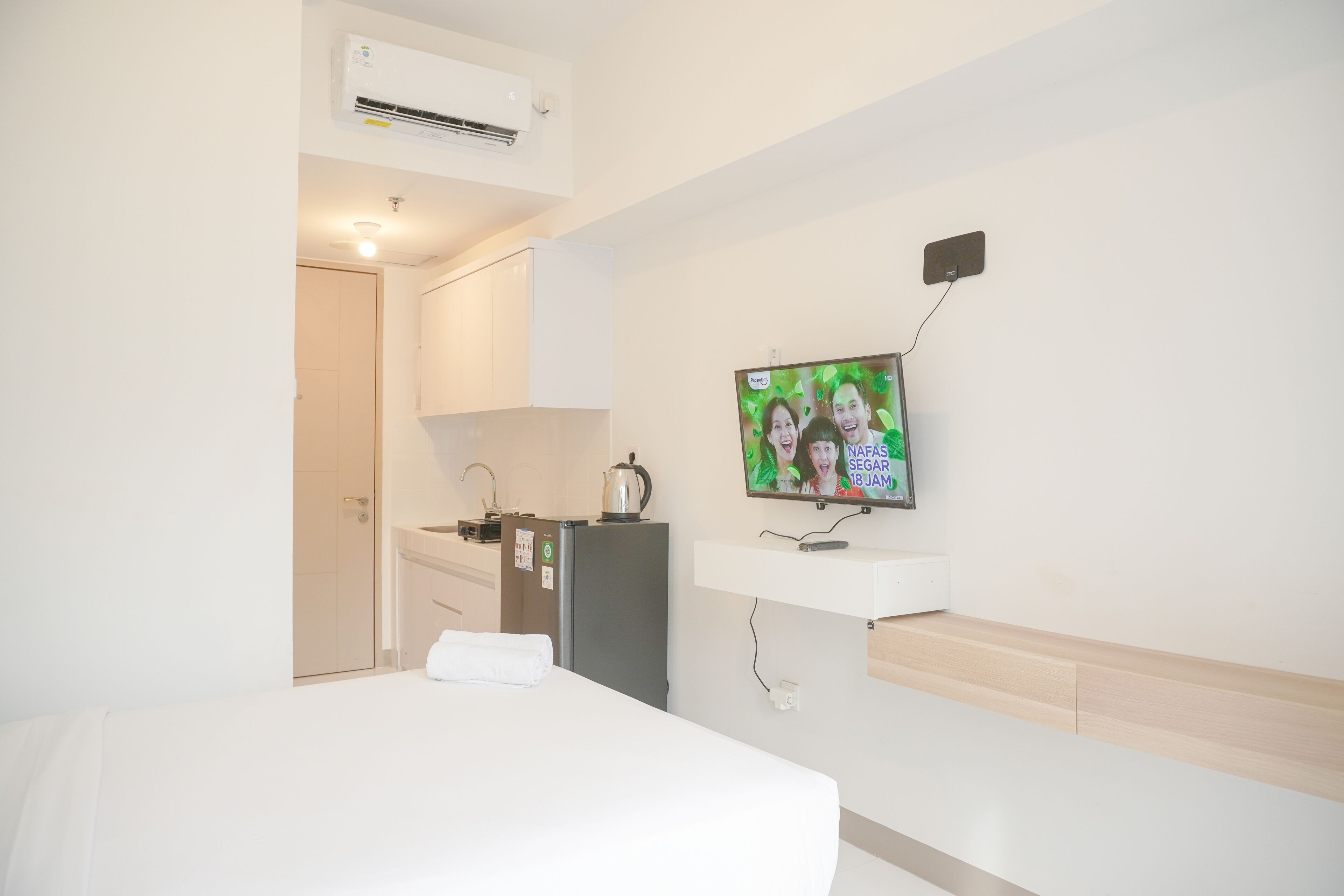 A hotel room with a bed, a TV mounted on the wall, a small kitchenette, and an air conditioning unit.