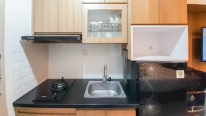 A compact kitchen with wooden cabinets, a black countertop, a stainless steel sink, and a black refrigerator with a green sticker. - Relaxing Studio At Taman Melati Sinduadi Apartment (Pogung Lor)