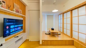 A modern living room with a flat-screen TV on a white entertainment unit, a wooden table with a small rug, and a sliding shoji screen door. - Spacious And Homey 1Br Tokyo Riverside Pik 2 Apartment (Teluknaga)