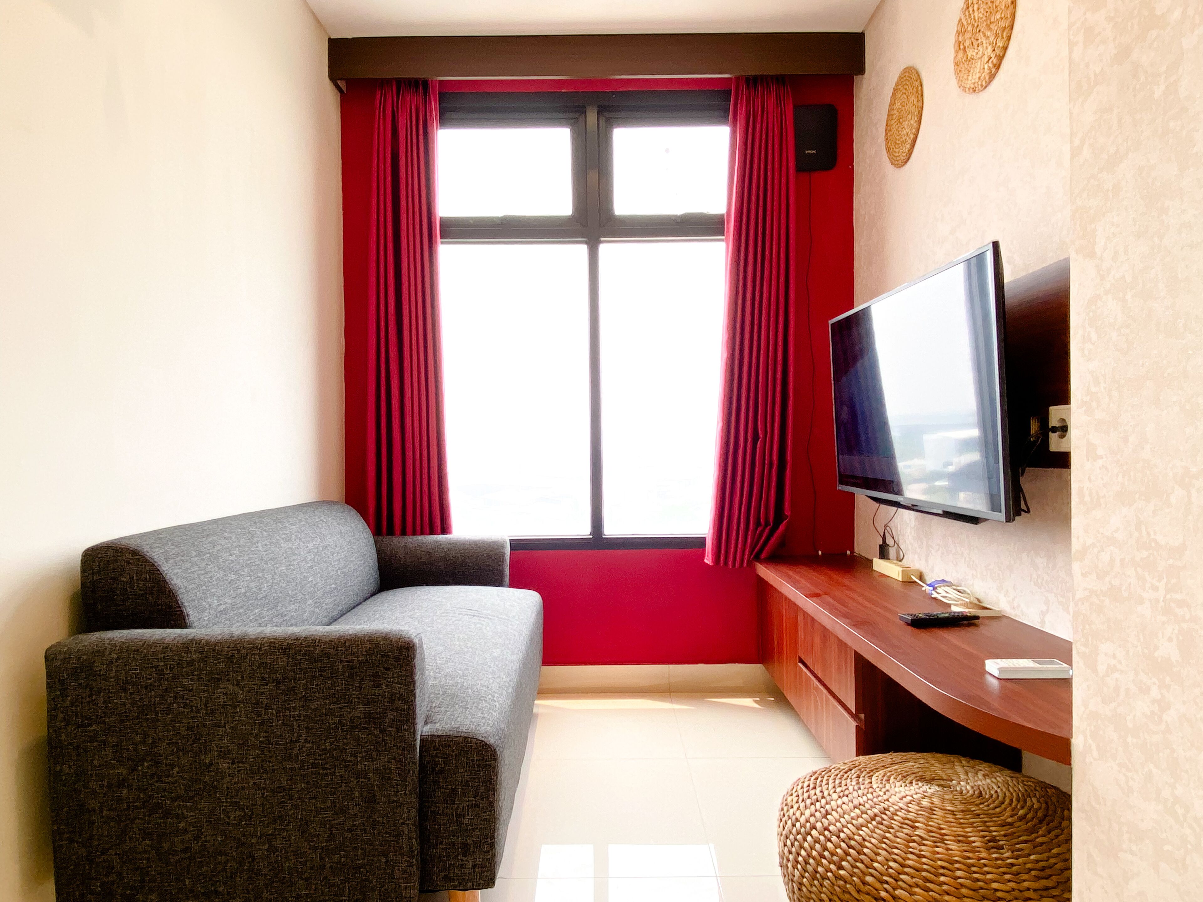 A room with a grey sofa, a flat-screen TV mounted on the wall, a wooden desk, and a wicker ottoman.