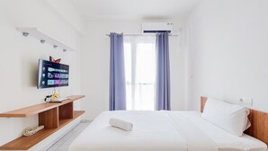 A modern hotel room with a bed, a TV mounted on a wooden shelf, and a window with curtains. - Warm Studio At 38Th Floor Sky House Alam Sutera Apartment (Tangerang)