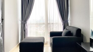 Un salon moderne avec un canapé et un pouf, de grandes fenêtres avec des rideaux et une vue sur le paysage urbain.