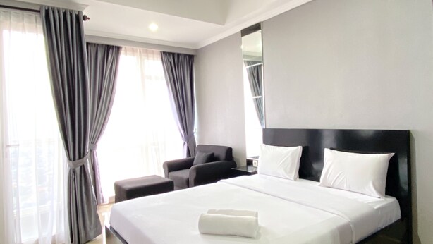 Une chambre d’hôtel moderne dotée d’un grand lit, d’un coin salon avec un fauteuil et un pouf, et d’une grande fenêtre avec des rideaux.