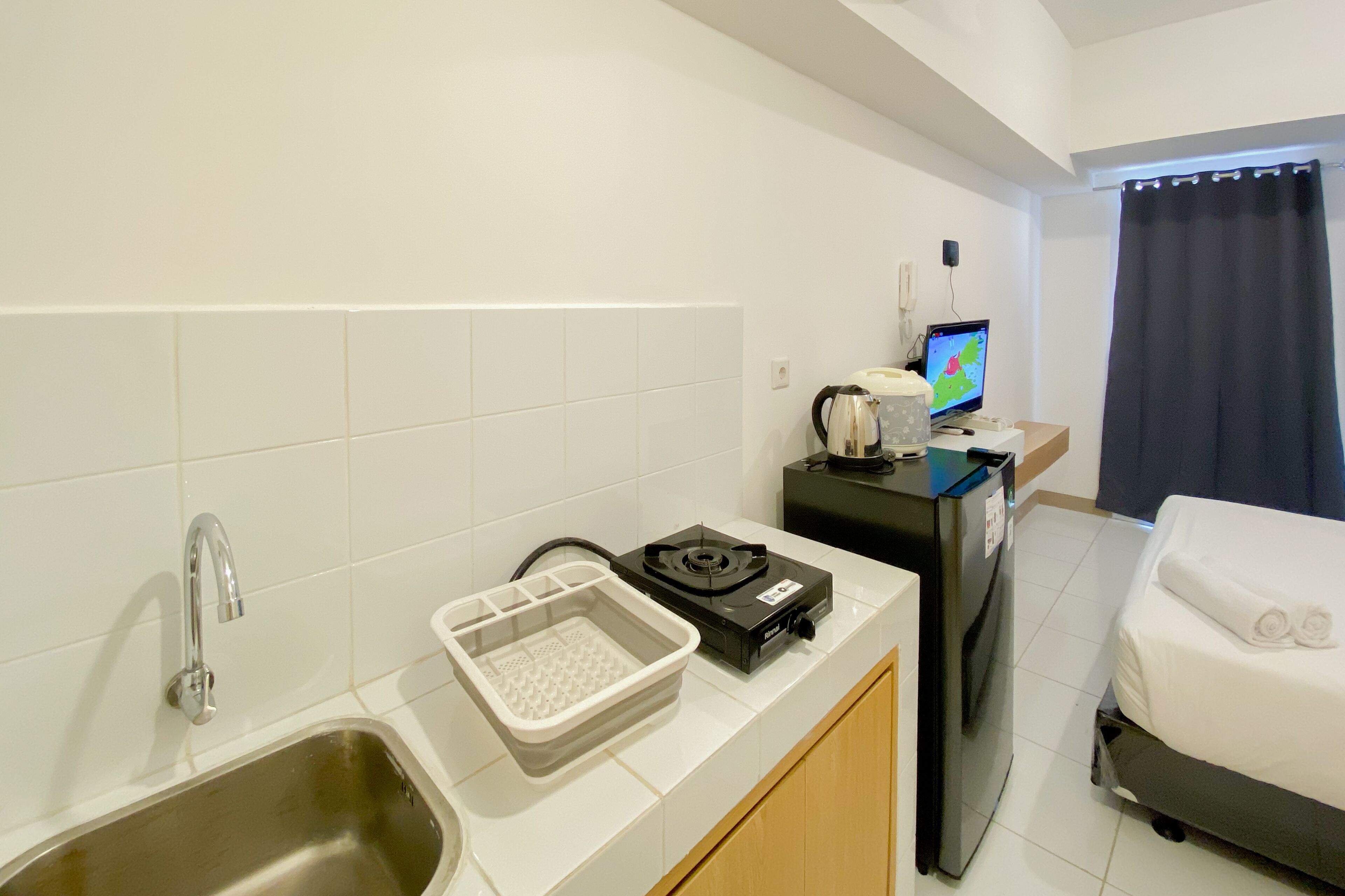 A compact hotel room with a kitchenette, a bed with neatly folded towels, a small desk with a computer, and a window with a curtain.